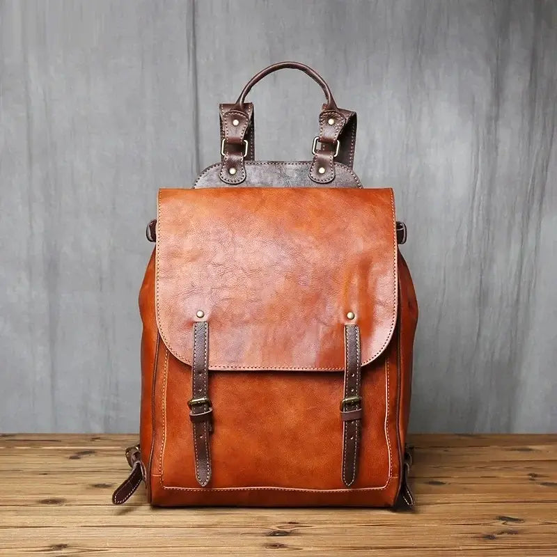 Sac à dos homme cuir vintage posé debout sur une table en bois, avec rabat et boucles en cuir foncé devant un rideau gris.
