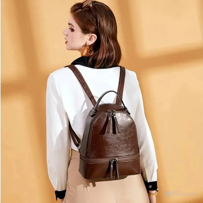 Sac à dos pour femme marron foncé porté par une femme en chemisier blanc et pantalon beige, lumière naturelle sur fond orangé.