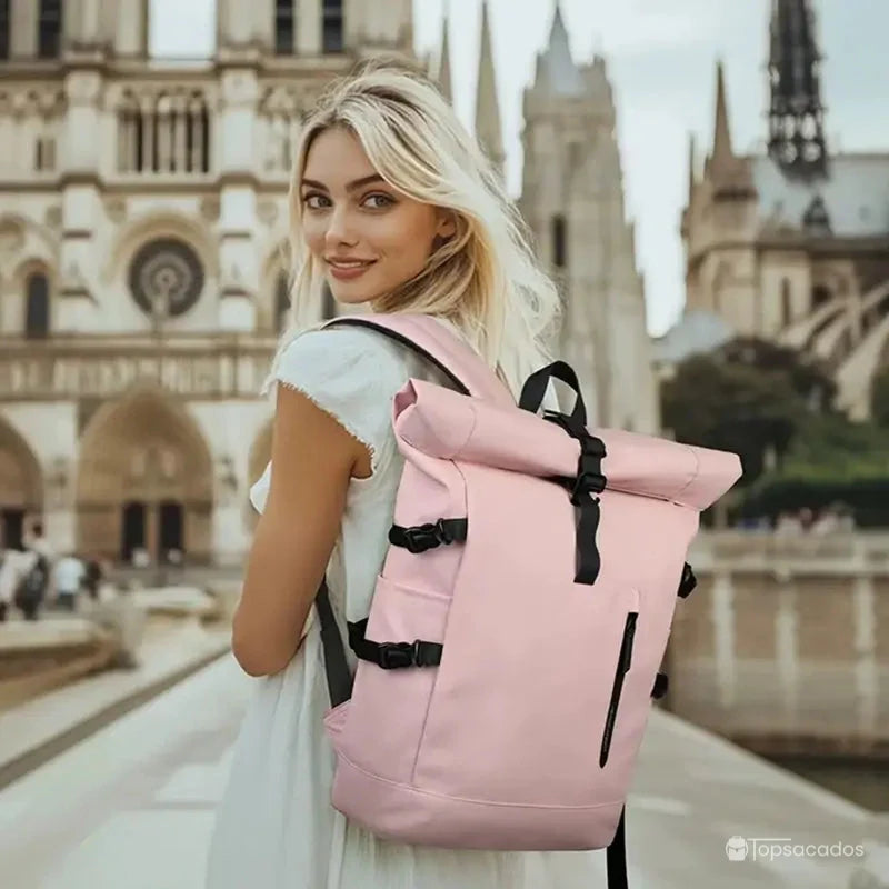 Sac a dos Roll Top pour femme rose porté par une jeune femme blonde en robe claire, marchant dans un centre-ville historique sous une lumière naturelle.